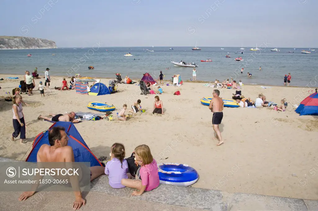 England, Dorset, Swanage Bay, A Man Sat With Small Children Looking Out Towards Busy Sandy Beach With Sunbathers On The Sand And Swimming In The Sea