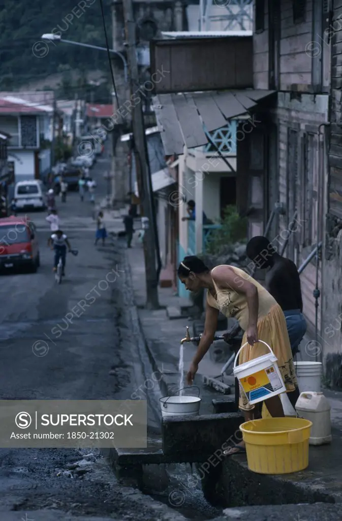 St Lucia, Soufriere, Man And Woman Collecting Water From Standpipe In Narrow Street With Open Gutter.