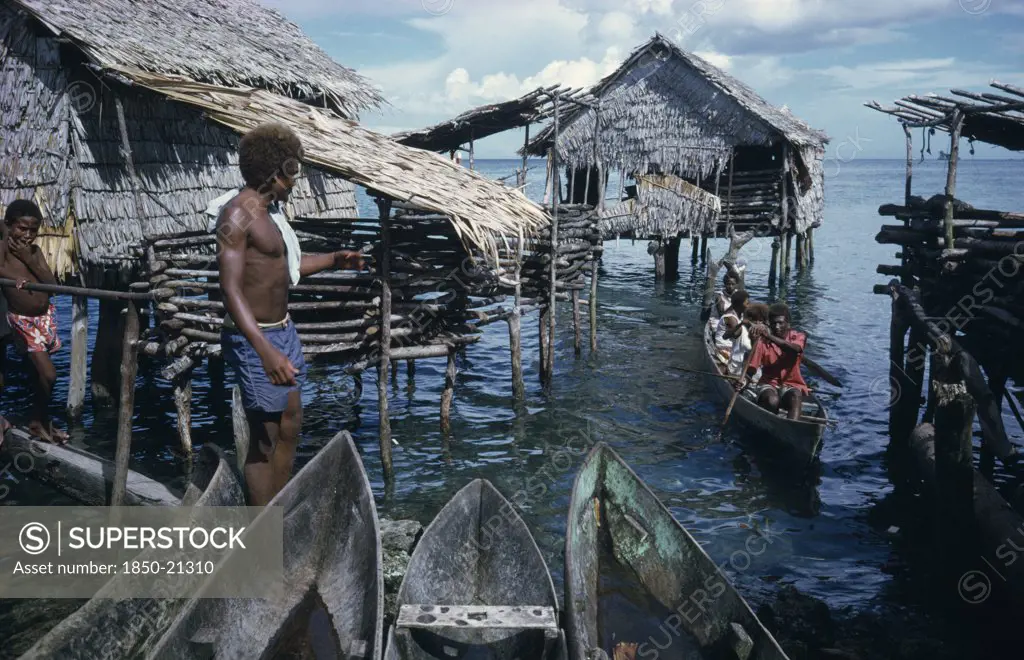 Pacific Islands, Melanesia, Solomon Islands, Thatched Stilt Houses On Artificial Island With Man And Child On Wooden Bridge And Approaching Canoe.  Part View Of Wooden Canoes In Immediate Foreground.