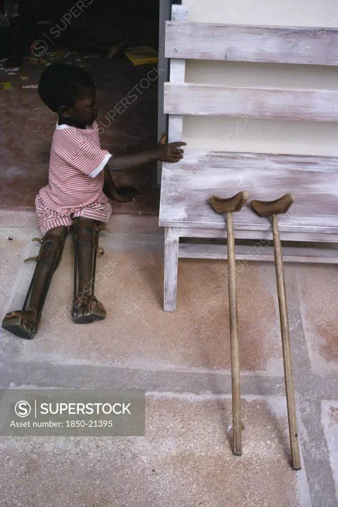 Tanzania, Medical, Child With Both Legs In Braces Sitting In Open Doorway Of Polio Unit Beside Pair Of Crutches Leaning Against Outside Wall.