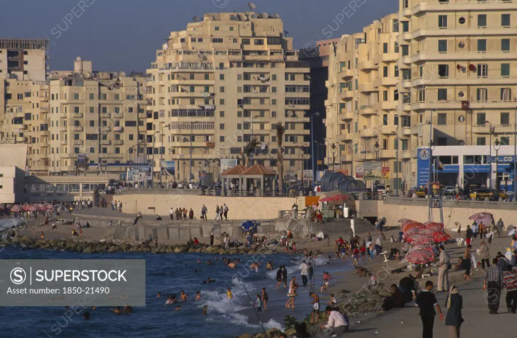 Egypt, Nile Delta, Alexandria, 'Corniche Waterfront Seen In Golden Light. Busy Beach With People Swimming In The Sea, Fishing From The Waters Edge And Walking On The Promenade. Overlooked By Tall Buildings.'