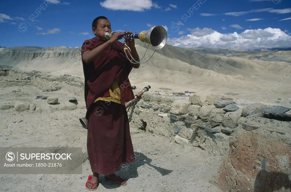 Nepal, Mustang, Namgyal Gompa, 'Tibetan Buddhist Monk Blowing Long, Decorated Horn On Monastery Roof To Summon The Faithful To Prayer.  Barren, Mountainous Landscape Behind. Rtnd 2 Vkb 15/5/2009'