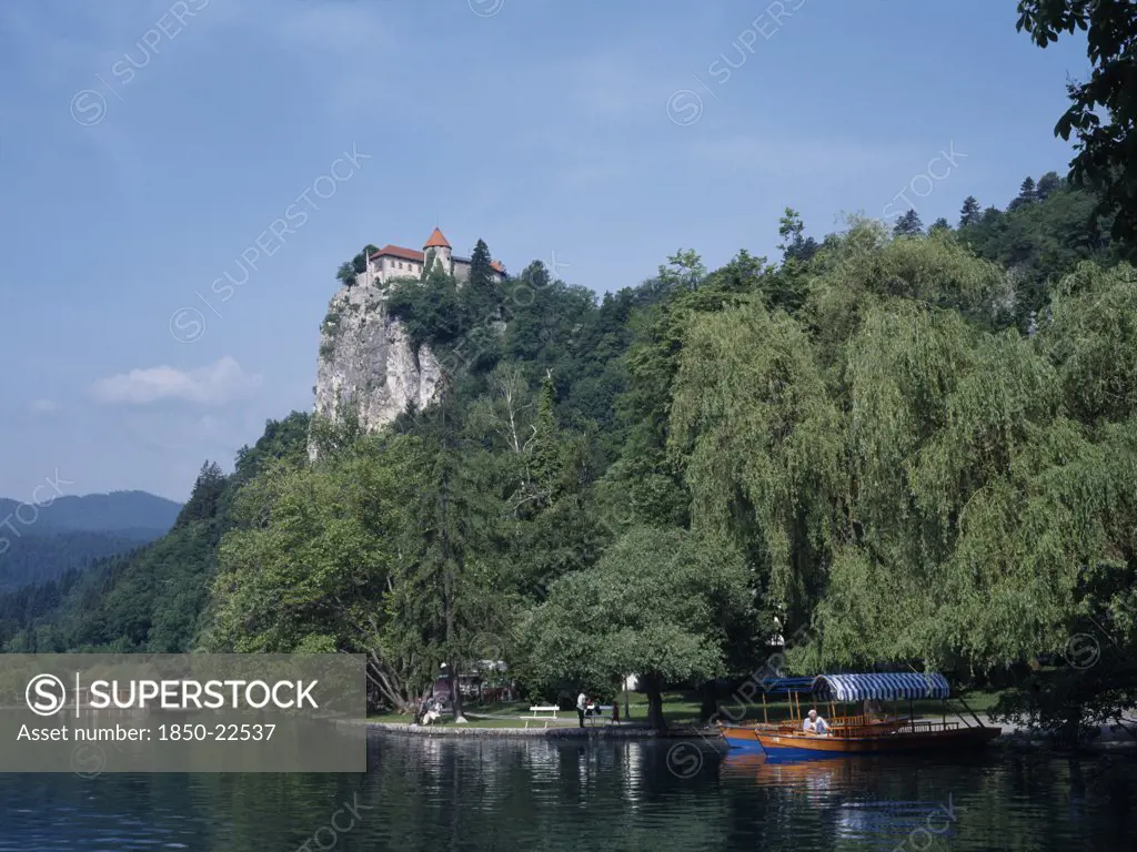 Slovenia, Lake Bled, Tourist Boats On Lake Bled With Castle Partly Seen Amongst Trees High Above.  Few People On Lake Shore Below.