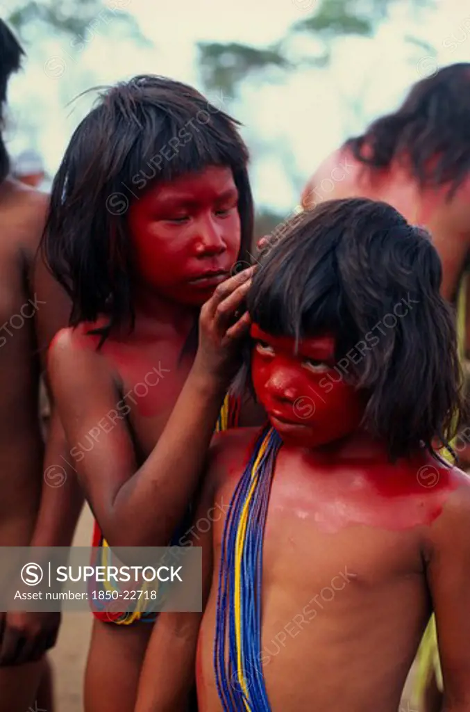 Brazil, Mato Grosso, Indigenous Park Of The Xingu, Young Panara Girls Applying Red Karajuru Face And Body Paint In Preparation For Dance.  Both Wearing Multiple Strands Of Blue Red And Yellow Beads Across Upper Body. Formally Known As Kreen-Akrore  Krenhakarore  Krenakore  Krenakarore  Amazon American Brasil Brazilian Indegent Kids Kreen Akore Latin America Latino South America Immature