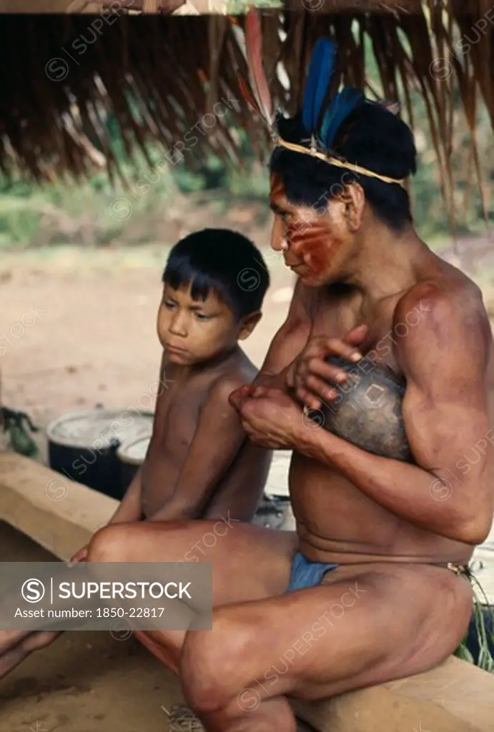 Colombia, North West Amazon, Vaupes, Maku Hunter  Face Painted With Red Achiote Marks And Wearing  Macaw Feather Crown In Preparation For Dance  Sits Beside Young Boy. Indigenous Tribe Indian Nomadic American Colombian Columbia Hispanic Indegent Kids Latin America Latino Male Men Guy South America Vaupes Immature Male Man Guy Vaupes