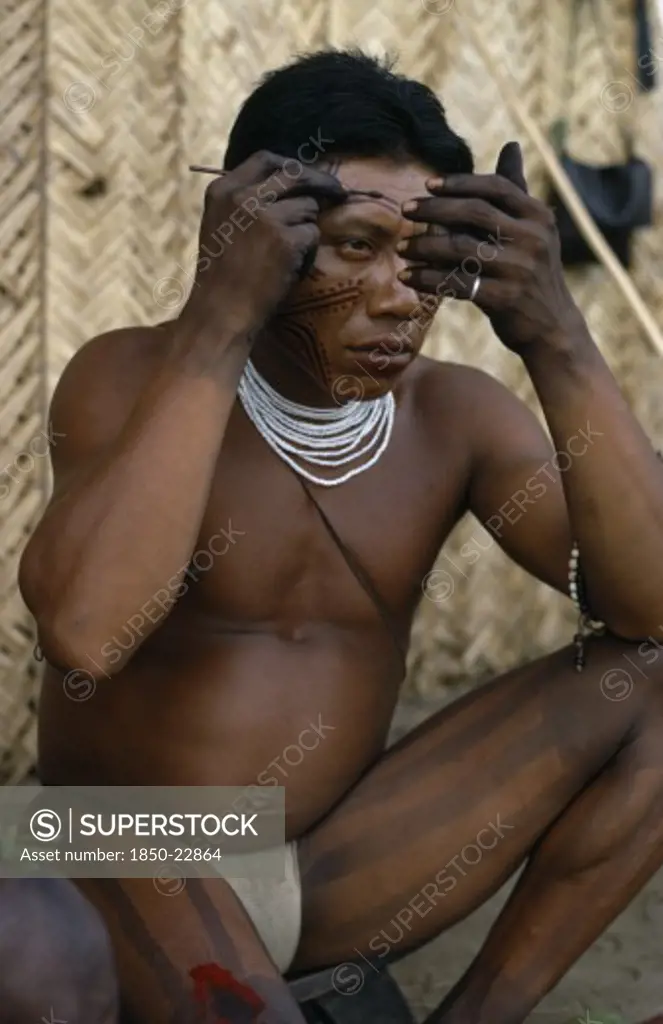Colombia, North West Amazon, Tukano Indigenous People, 'Barasana Man Looking In Mirror To Apply Red Achiote Facial Paint,  Thick Dark Purple We Leaf Juice Vertical Stripes Painted On Legs  Wearing Multi Stranded White Glass Bead Necklace.  Tukano Sedentary Indian Tribe North Western Amazonia Body Decoration American Colombian Columbia Hispanic Indegent Latin America Latino Male Men Guy South America Tukano  '