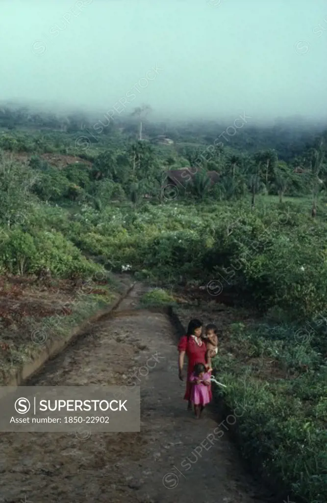 Colombia, North West Amazon, Tukano Indigenous People, Makuna Mother Carrying Baby And With Young Daughter In Front Of Her Walking Between Malocas Along Unmade Road Through Cleared And Cultivated Rainforest. Tukano  Makuna Indian North Western Amazonia Family American Babies Colombian Columbia Hispanic Indegent Kids Latin America Latino Mum Scenic South America Tukano