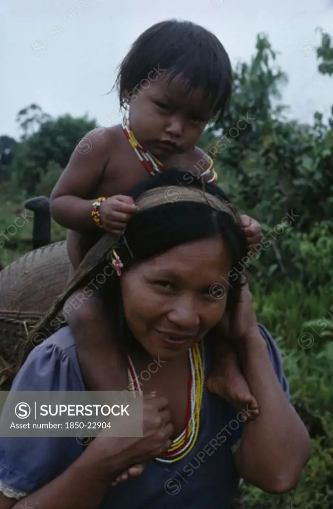 Colombia, North West Amazon, Tukano Indigenous People, Makuna Mother Carrying Her Baby Son On Her Shoulders And Basket On Her Back Supported By Head Strap.  Both  Wearing Necklaces Of White Red And Yellow Glass Beads. Tukano  Makuna Indian North Western Amazonia Family American Babies Colombian Columbia Hispanic Indegent Kids Latin America Latino Mum South America Tukano