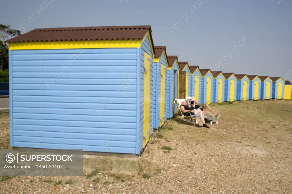England, West Sussex, Littlehampton, A Crescent Of Blue And Yellow Beach Huts With A Couple Sunbathing On Loungers