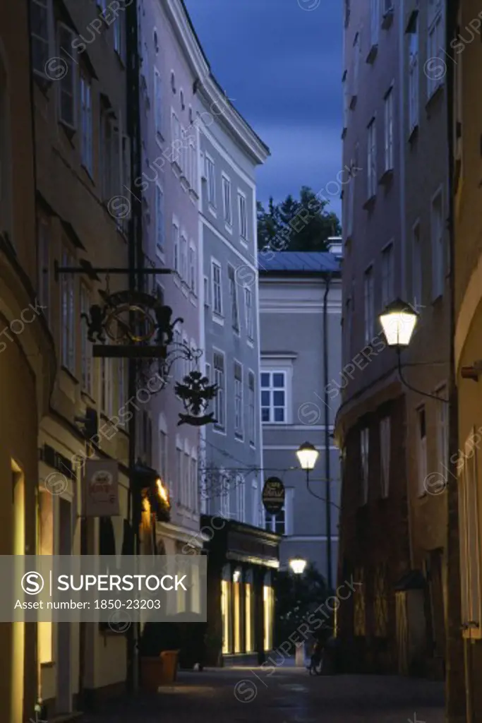 Austria, Salzburg, 'Narrow Street Of Judengasse At Night Lined By Tall, Grey, Pink And Cream Painted Buildings With Street Lamps Extending From Walls On Metal Brackets.'