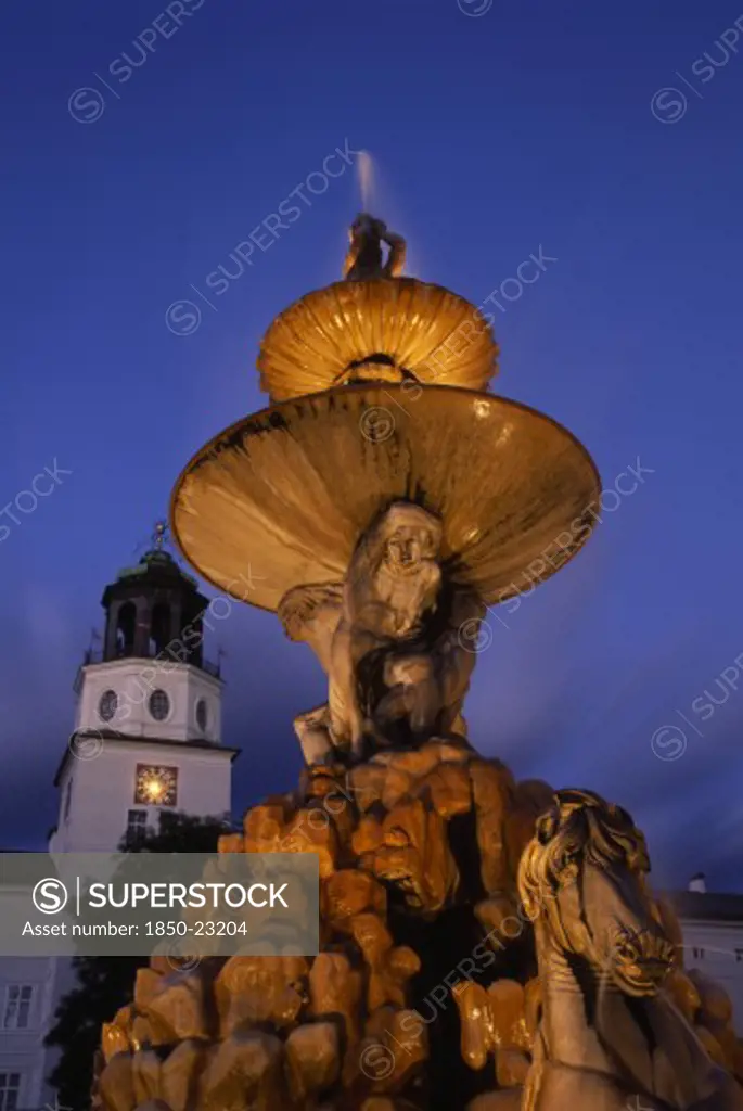Austria, Salzburg, Part View Of Baroque Fountain With Statues Of Horse And Contorted Figures And The Glockenspiel Carillon Tower Behind At Night.