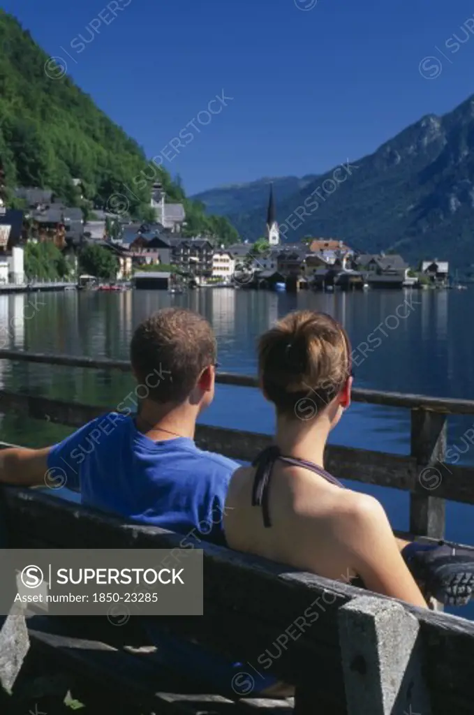Austria, Oberosterreich, Hallstatt, 'Young Couple Sitting On Wooden Bench Looking Out Across Hallstattersee Lake Towards Village Church, Houses And Boathouses With Mountain Backdrop.'