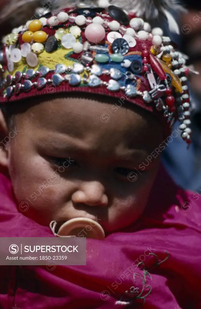 China, Xinjiang Province, Altai Region, Portrait Of Kazakh Baby Wearing Hat Covered With Silver Beads And Coloured Buttons.