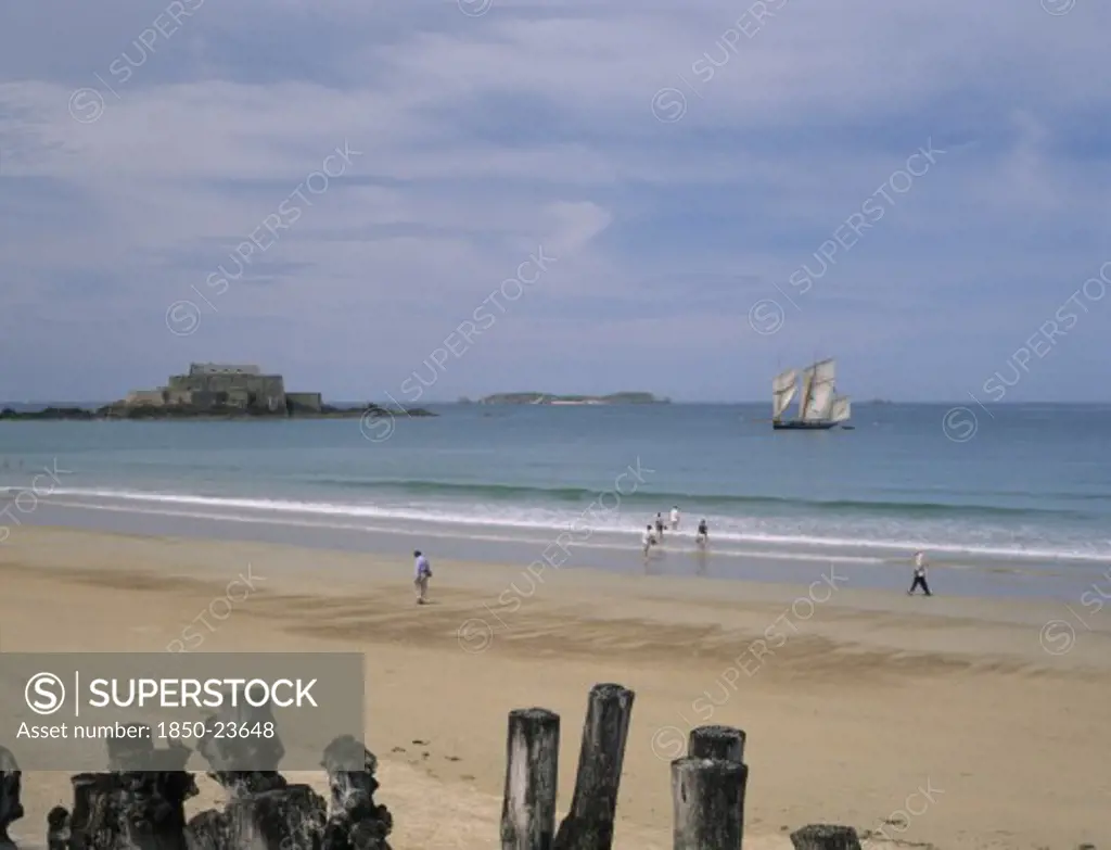 France, Brittany, St Malo, Le Sillon Beach And Fort National. People Walking Along Golden Sand With A Sail Boat On Seen On Water