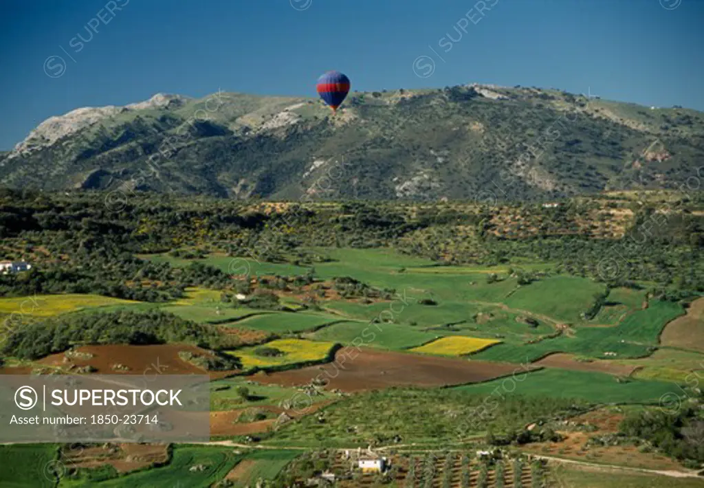 Spain, Andalucia, Malaga , A Hot Air Balloon Flying Over Green Valley With Patchwork Fields Towards Mountain Range