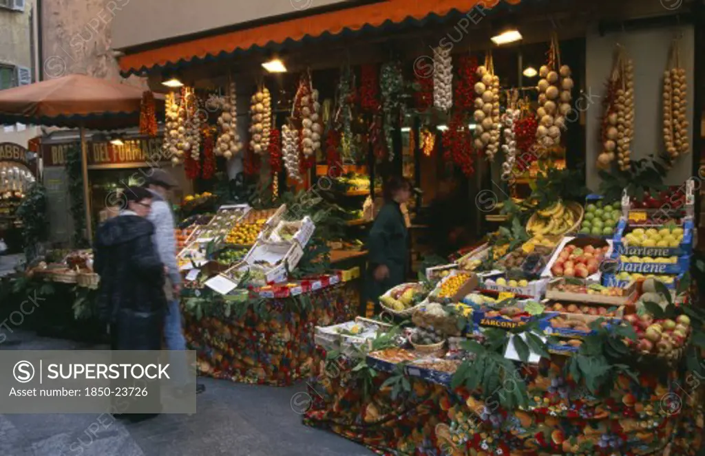 Switzerland, Ticino, Lugano, Member Of Staff And Customers Outside Fruit And Vegetable Stall With Pavement Display And Strings Of Onions And Garlic Hanging From Rack Above Doorway.