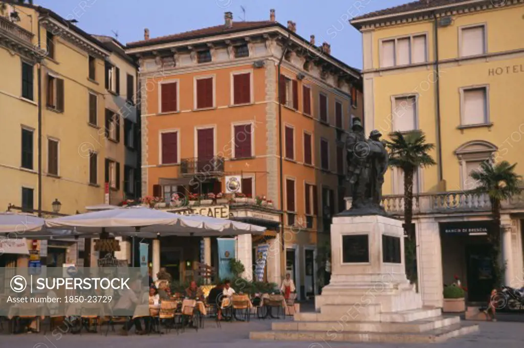 Italy, Lombardy, Salo, 'People Sitting At Outside Cafe Tables In Piazza Of Town Beside Lake Garda. Facades Of Buildings, Bank And Hotel Painted Pale Yellow And Orange With Window Shutters.'