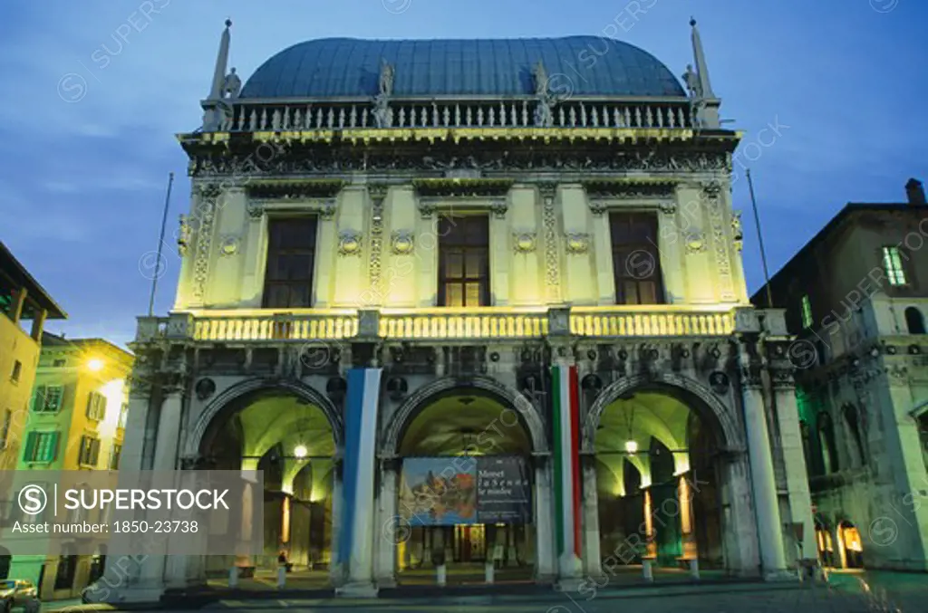 Italy, Lombardy, Brescia, 'Piazza Della Loggia.  Facade Of Loggia Or Town Hall With Arched Entrance And Colonnades, Balcony And Statues Illuminated At Night.  With Poster Advertising Monet Exhibition'