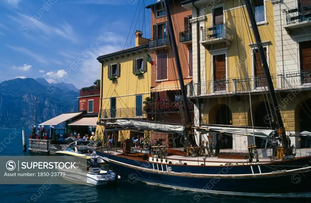 Italy, Veneto, Malcesine, 'Yellow, White And Terracotta Painted Facades Of Waterside Buildings With Moored Yacht And Motor Boats In Foreground.  People At Cafe At End Looking Out Across Lake '
