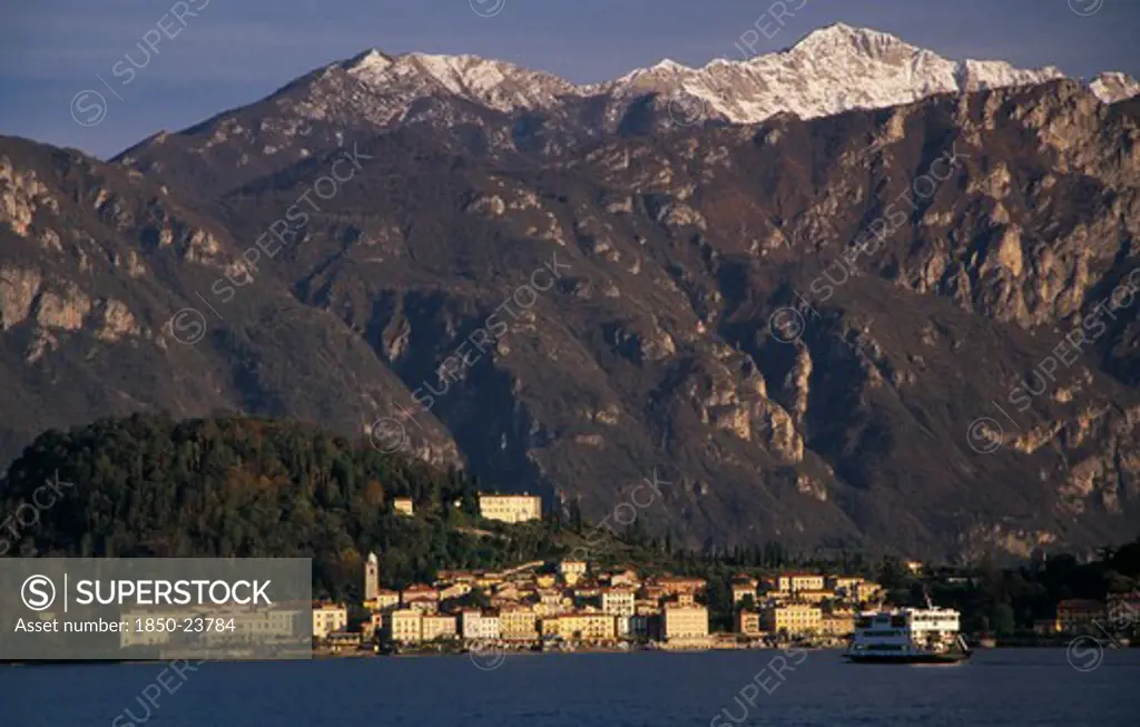 Italy, Lombardy, Lake Como, Ballagio. View Across Lake Como Towards Distant Town Of Ballagio Situated At Foot Of Tree Covered Hillside With Mountain Backdrop. Car Ferry Crossing Lake In Foreground.