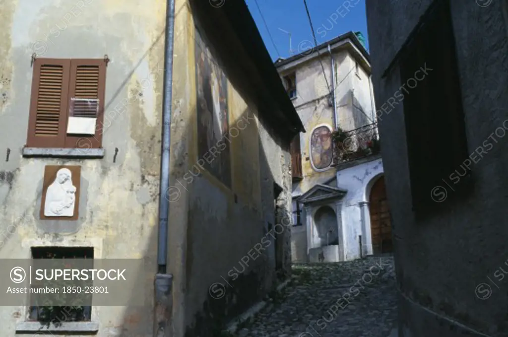 Italy, Lombardy, Arcumeggia, Narrow Cobbled Street With Fresco Decoration And Plaster Relief Of The Virgin And Child On The Exterior Walls Of The Buildings And Drinking Fountain At Top.