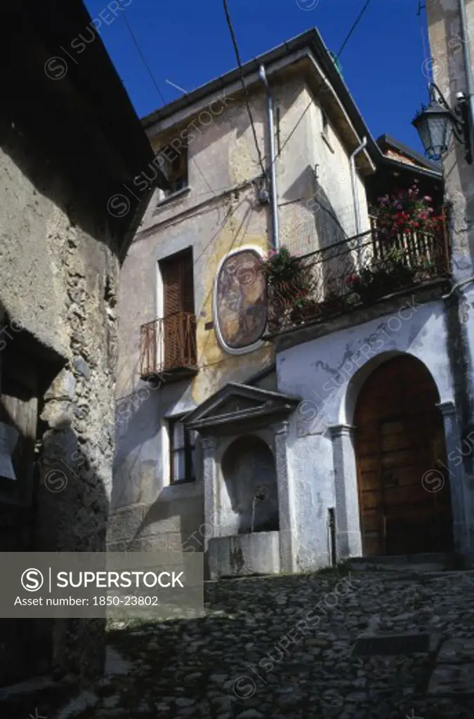 Italy, Lombardy, Arcumeggia, Narrow Cobbled Street With Fresco Decoration On Exterior Wall Of Building And Drinking Fountain Below Beside Arched Doorway With Balcony And Flowers Above.