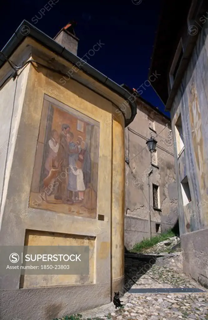 Italy, Lombardy, Arcumeggia, Narrow Cobbled Street With Fresco Decoration On Exterior Wall Of Corner Building.