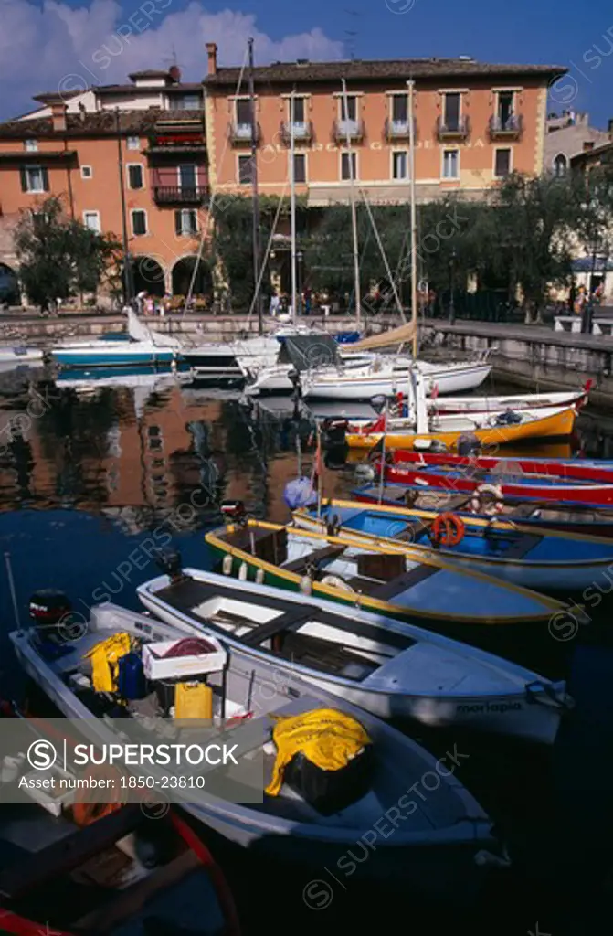 Italy, Veneto, Lake Garda, Torre Di Benaco.  Waterside Buildings Overlooking Harbour With Moored Boats Reflected In Water In The Foreground.