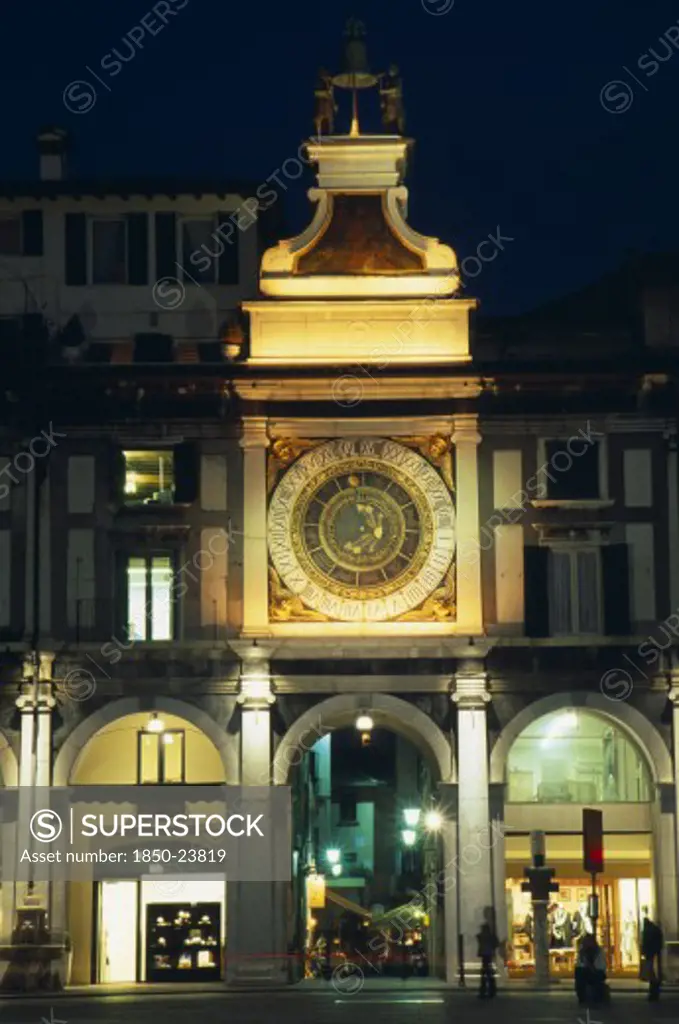 Italy, Lombardy, Brescia, 'Astronomical Clock In Piazza Della Loggia Illuminated At Night With Brightly Lit Shop Windows Framed By Archways Of Colonnade Below.  People, Street Lamps And Light Trail.'