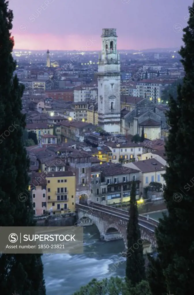Italy, Veneto, Verona, 'Cityscape At Sunset Showing Tiled Rooftops, Duomo Bell Tower And Bridge Over The Adige River Part Framed By Tall Coniferous Trees.  Pale Pink/Purple Sky And City Lights.'