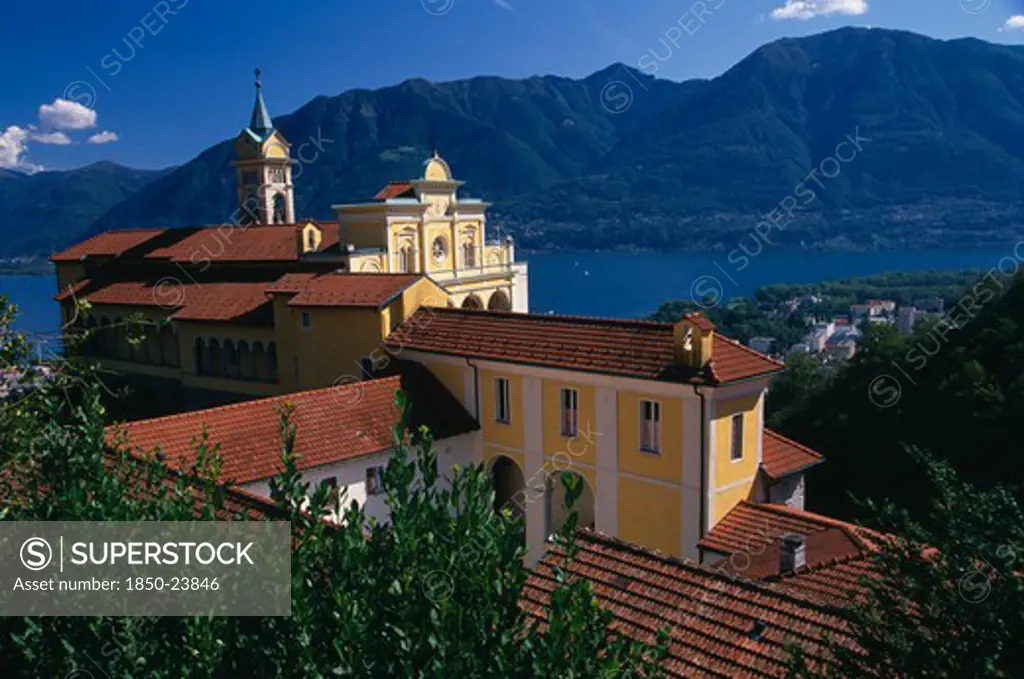 Switzerland, Ticino, Lake Maggiore, Locarno.  Tiled Rooftops And Bell Tower Of Yellow And White Painted Church Of Madonna Del Sasso With Lake And Mountains Beyond.