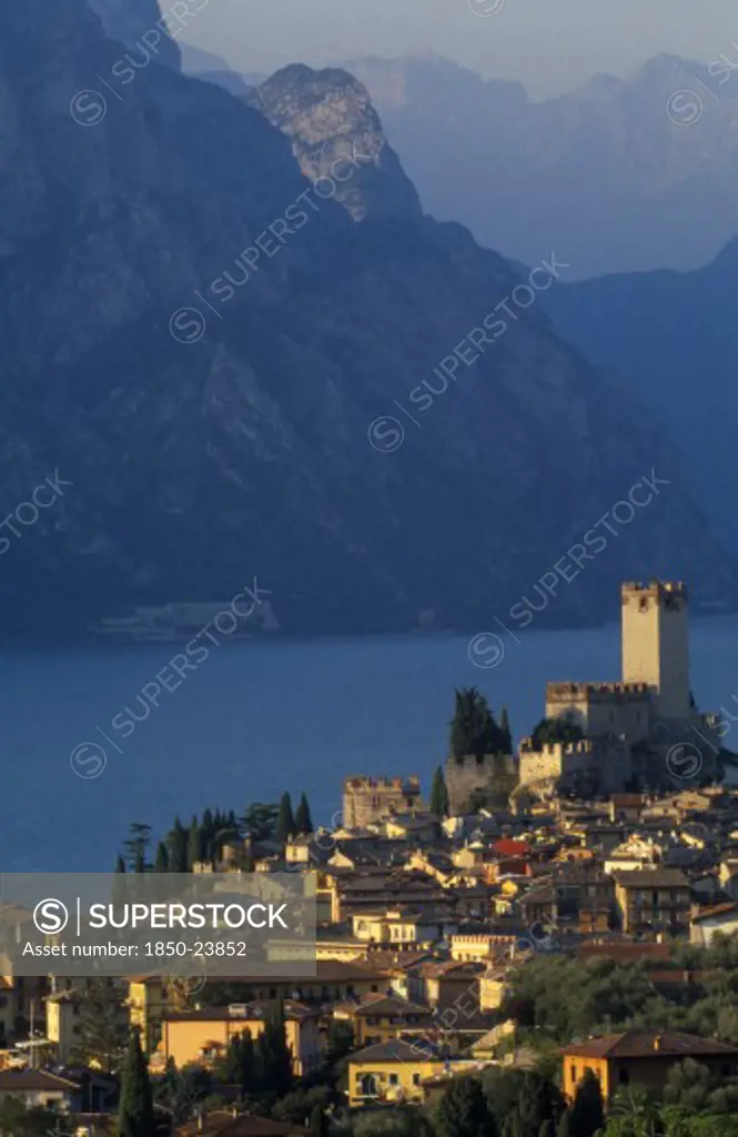 Italy, Veneto, Lake Garda, 'Malcesine.  View Across Town Rooftops Towards Castello Scaligero In Warm, Golden Light With Lake And Mountains Beyond.'