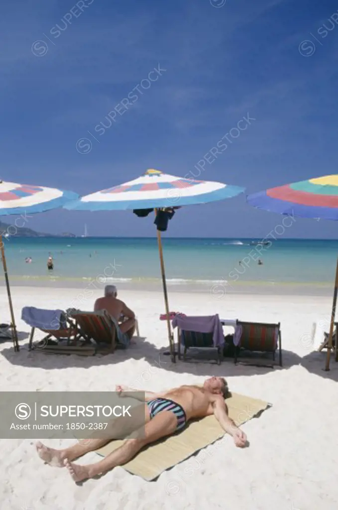 Thailand, Phuket, Patong Beach, Sun Bather Lying On The Sand In The Foreground With Row Of Umbrellas And Deck Chairs Behind And The Sea Beyond
