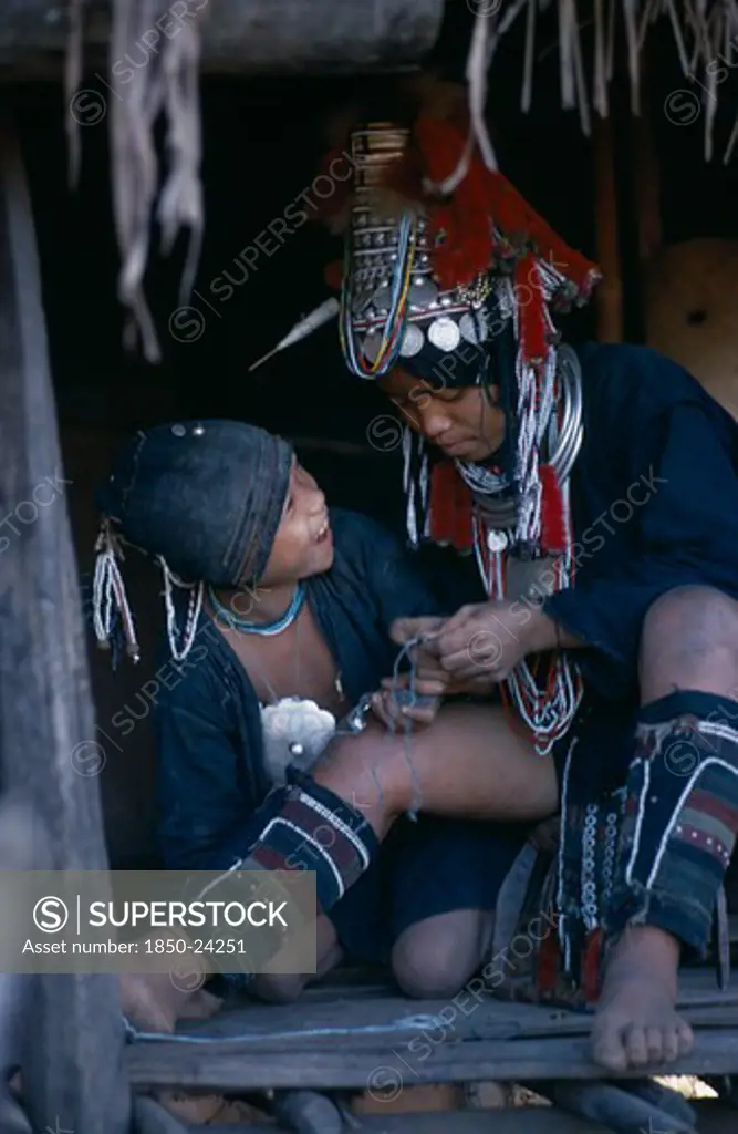 Burma, People, Akha Woman Wearing Traditional Decorated Silver Head-Dress And Leggings Sitting With Child In Hut Entrance.