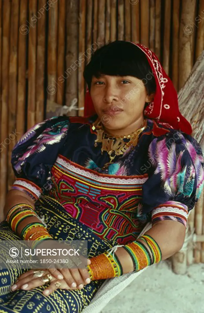 Panama, San Blas Islands, Kuna Indigenous Tribe, 'Portrait Of A Kuna Indian Woman In Hammock  With Gold Jewelry Including Gold Nose Ring And Rings On Fingers, Bead Design Arm Bands, Wearing Brightly Coloured Layered Applique Traditional Mola With Fine Bird Design  Beads On Arms And A Gold Nose Ring. Cuna'
