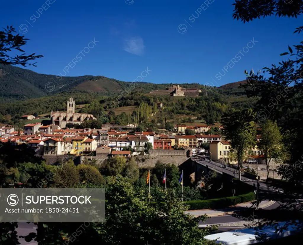 France, Languedoc-Roussillon, Pyrenees-Orientales, Prats-De-Mollo-La-Preste.  Mountain Town With Typical Painted Houses With Red Tiled Rooftops And Fort Lagarde On Hillside Behind.  Road And Tree Tops In The Foreground.