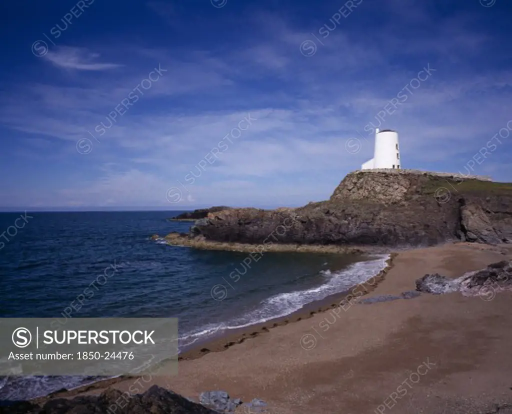 Wales, Isle Of Anglesey, Llanddwyn Island, 'Curving, Empty Beach Overlooked By Old Lighthouse Standing On Rocks Extending Out To Sea.  Blue Sky And Windswept Clouds Above.'