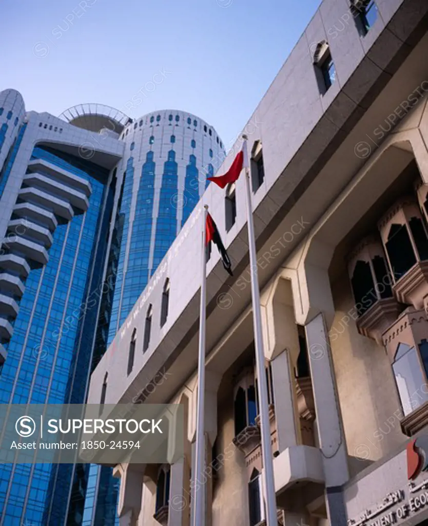 Uae, Dubai, 'Two Flag Poles In Front Of Contrasting Styles Of Architecture, Modern, Mainly Glass Skyscraper Behind Facade Of Building In More Traditional Style.'