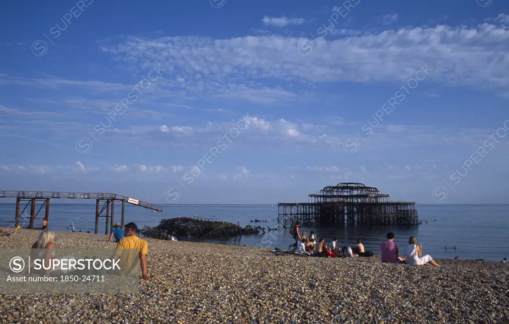 England, East Sussex, Brighton, Groups Of People Sitting On The Beach By The Ruined West Pier In Late Afternoon Sunshine.