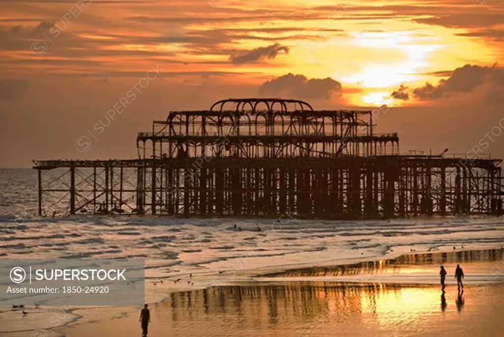 England, East Sussex, Brighton, 'People Walking Along The Wet Sand At Low Tide At Sunset, With A Dramatic Skyline, In Front Of The Remains Of The West Pier'