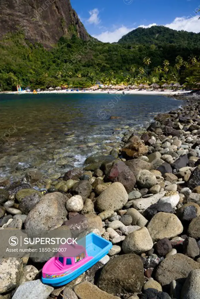 West Indies, St Lucia, Soufriere , Val Des Pitons The White Sand Beach At The Jalousie Plantation Resort Hotel With The Volcanic Plug Of Petit Piton Beyond And Tourists On Sunbeds Beneath Palapa Sun Shades With A Childs Plastic Boat Marooned On Rocks In The Foreground