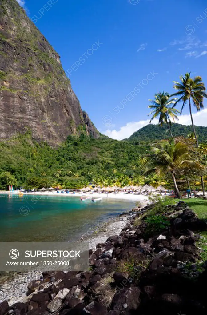 West Indies, St Lucia, Soufriere , Val Des Pitons The White Sand Beach At The Jalousie Plantation Resort Hotel With The Volcanic Plug Of Petit Piton Beyond And Tourists On Sunbeds Beneath Palapa Sun Shades