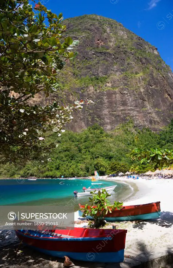 West Indies, St Lucia, Soufriere , Val Des Pitons The White Sand Beach At The Jalousie Plantation Resort Hotel With The Volcanic Plug Of Petit Piton Beyond And Tourists On Sunbeds Beneath Palapa Sun Shades With Colourful Fishing Boats Under A Tree In The Foreground