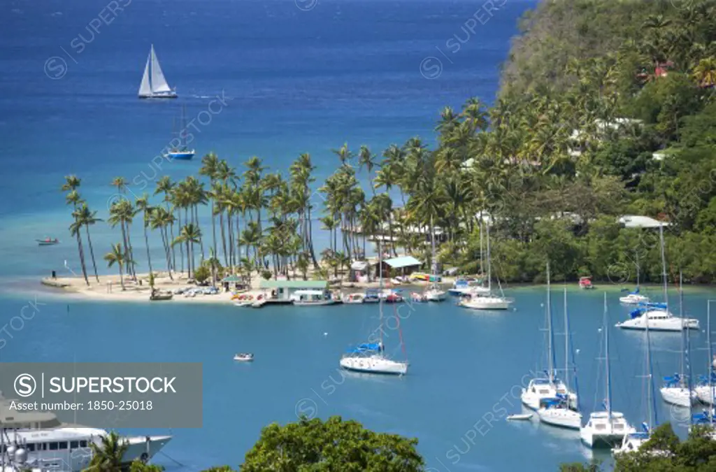 West Indies, St Lucia, Castries , Marigot Bay The Harbour With Yachts At Anchor And A Yacht Sailing Out At Sea Beyond The Small Coconut Palm Tree Lined Beach Of The Marigot Beach Club Sitting At The Entrance