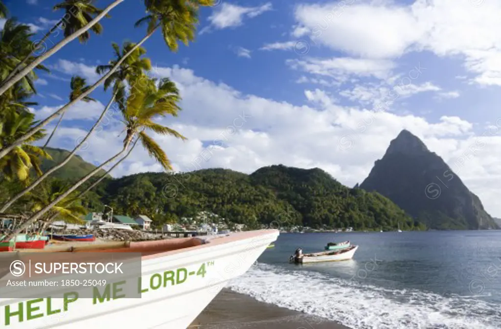 West Indies, St Lucia, Soufriere, Fishing Boats On The Beach Lined With Coconut Palm Trees With The Town And The Volcanic Plug Mountain Of Petit Piton Beyond. A Fishing Boat In The Foreground With The Words Help Me Lord Written On The Bow