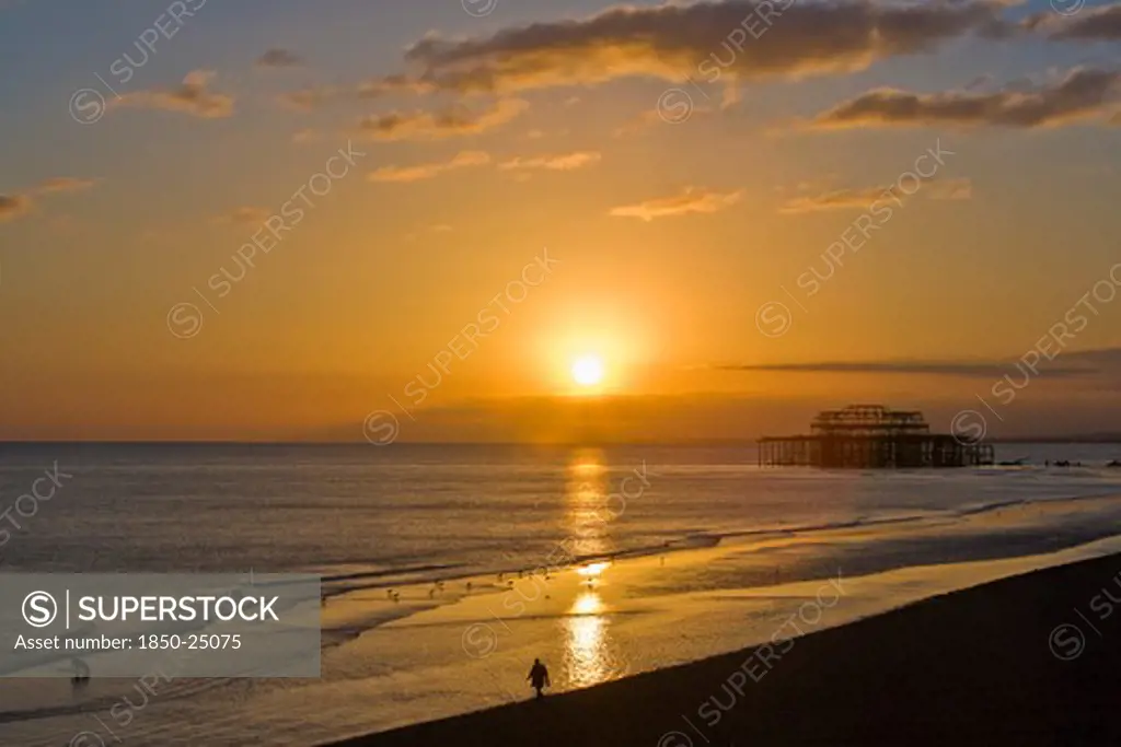 England, East Sussex, Brighton, Dramatic Orangy-Red Sunset Silhouetting Remains Of The West Pier With Dog And Walker On The Beach In The Foreground
