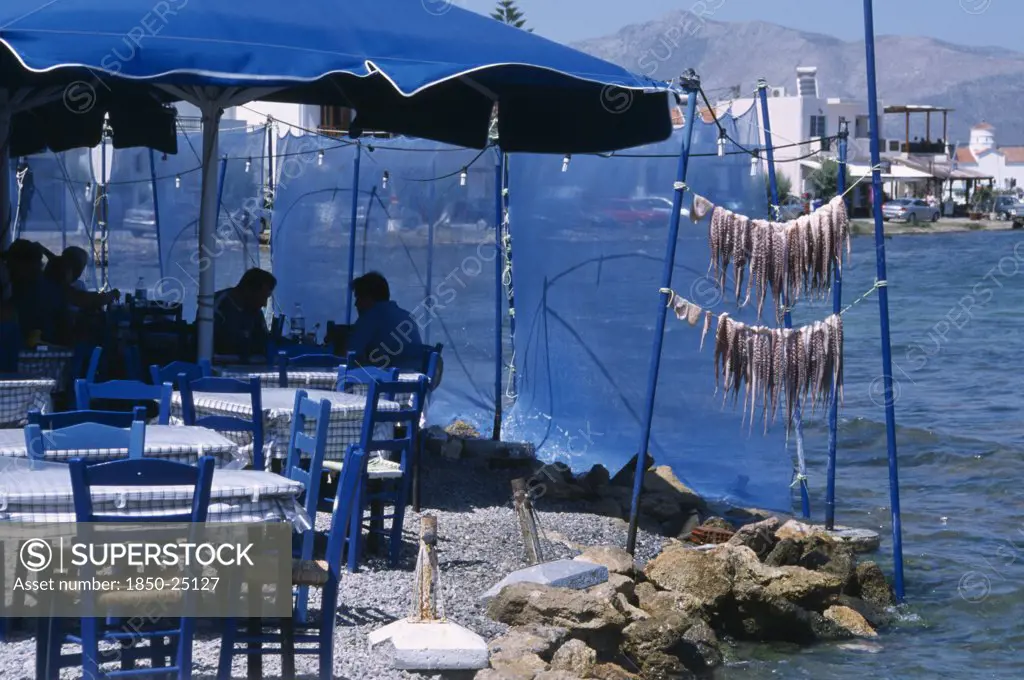 Greece, Peloponnese, Elafonisos , Restaurant Outside Seating With Blue Table And Chairs Next To Waters Edge With A Fresh Catch Of Seafood Hanging From Poles