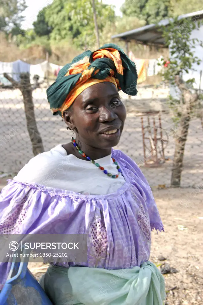 Gambia, Western Gambia, Tanji, Tanji Village.  Three-Quarter Portrait Of Woman Wearing Traditional Head Tie Or Scarf And Bead Necklace Posing In Front Of Her Home Compound.