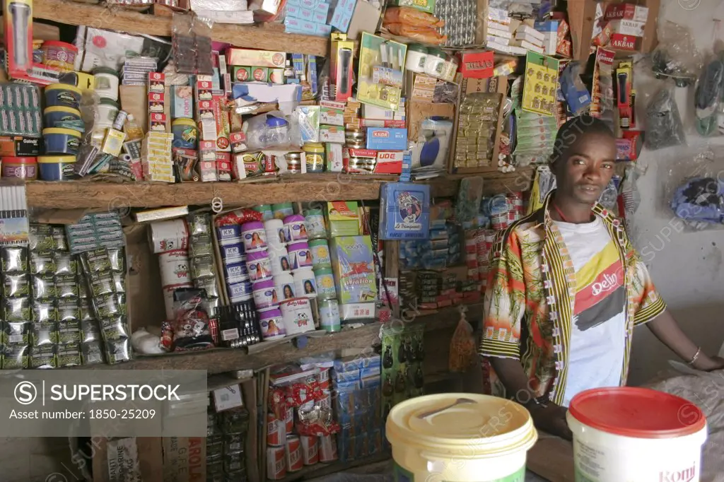 Gambia, Western Gambia, Tanji, 'Tanji Village.  Young, Male Owner Of A Grocery Shop Standing Behind Wooden Counter In Front Of Shelves With Display Of Various Products. '