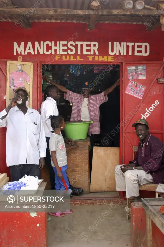 Gambia, Atlantic Coast, Banjul, 'Shop In Gambia'S Capital Banjul Painted Red And White, The Colours Of The Manchester United Football Strip And Decorated With Slogans And Posters With Group Of African Supporters Of The Team Gathered Outside And In Doorway.'
